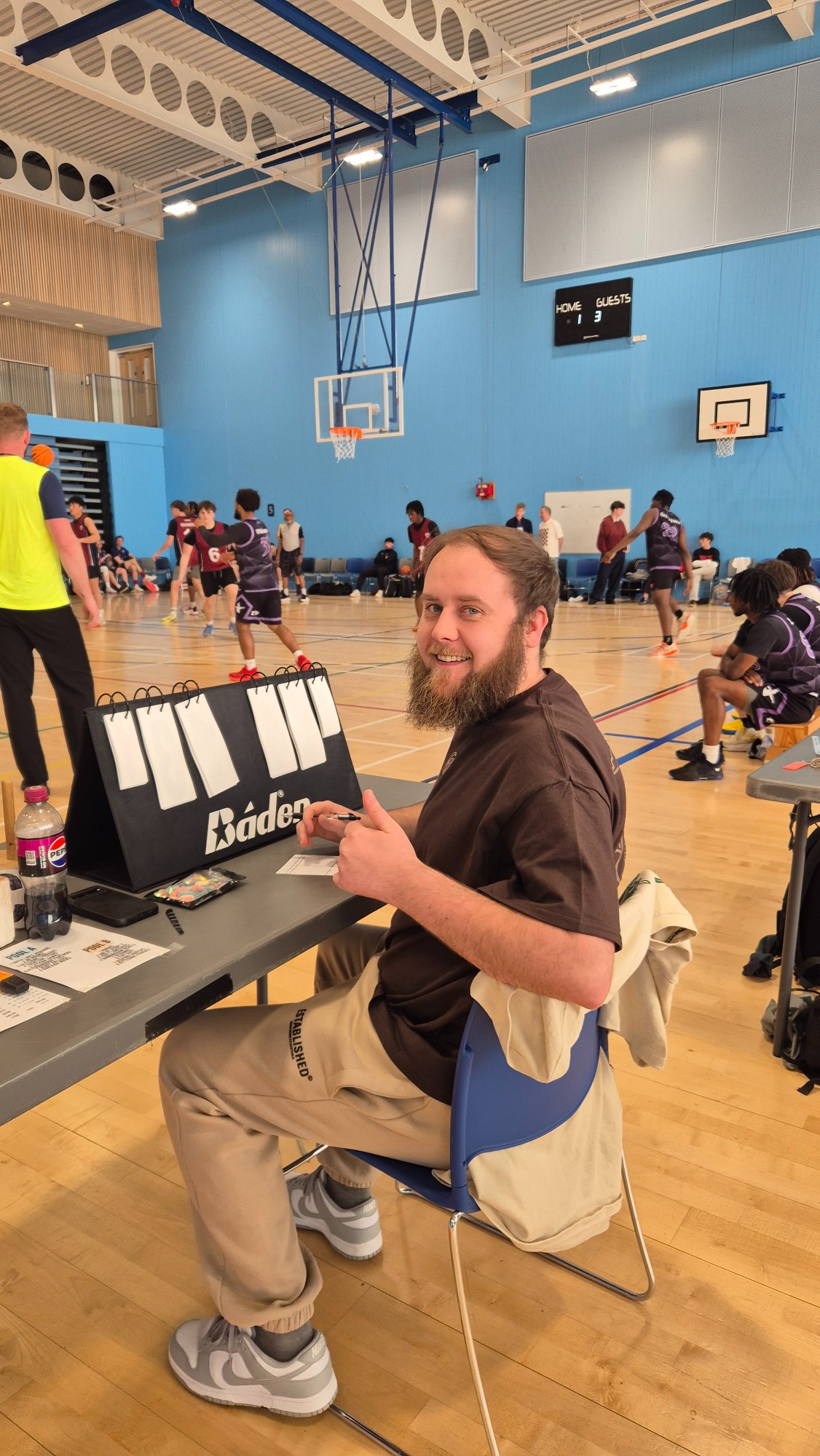 A man sitting at a table, smiling and giving a thumbs up, amidst a basketball court scene.