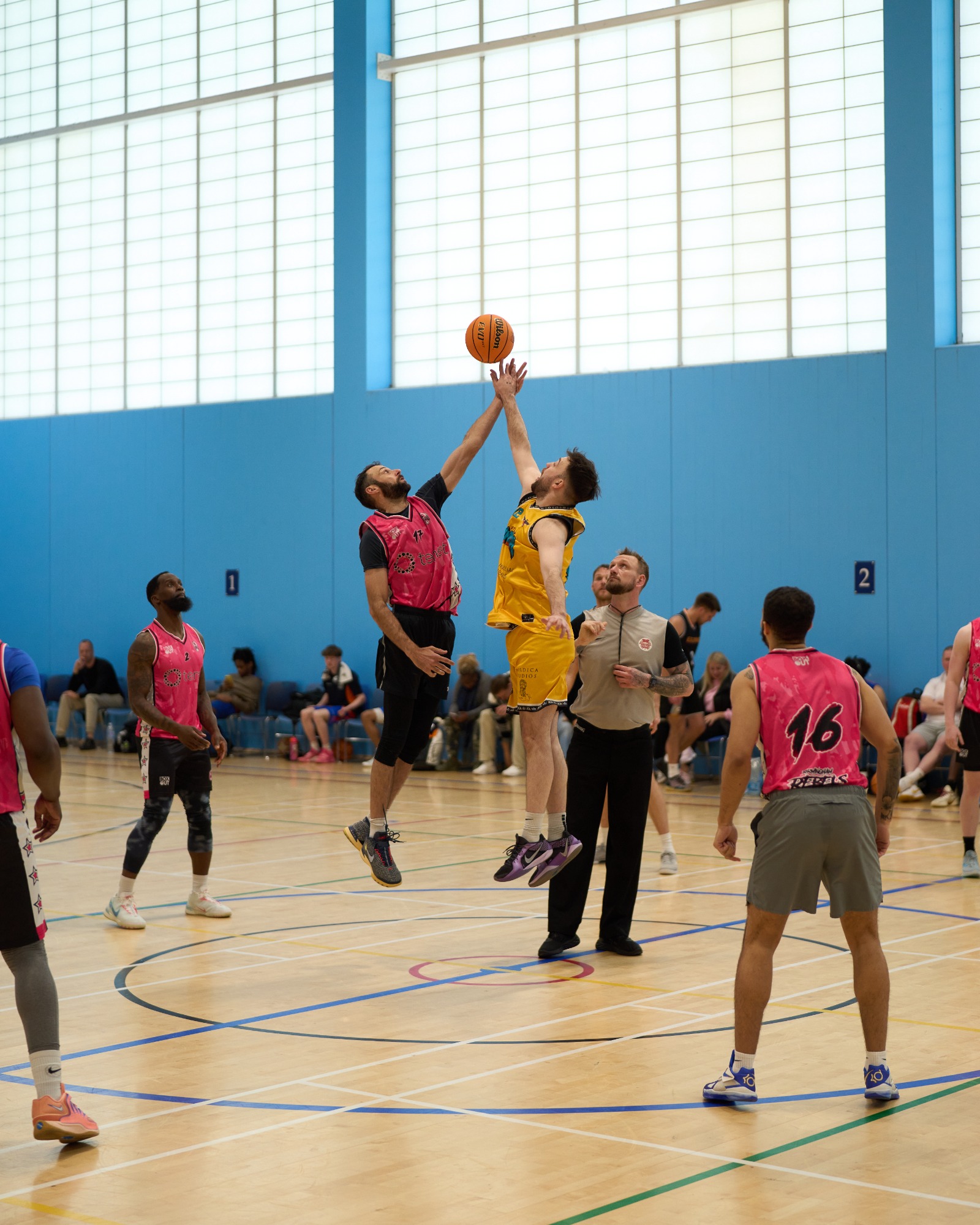 Two basketball players jump for the ball at the start of a game, surrounded by teammates and spectators.