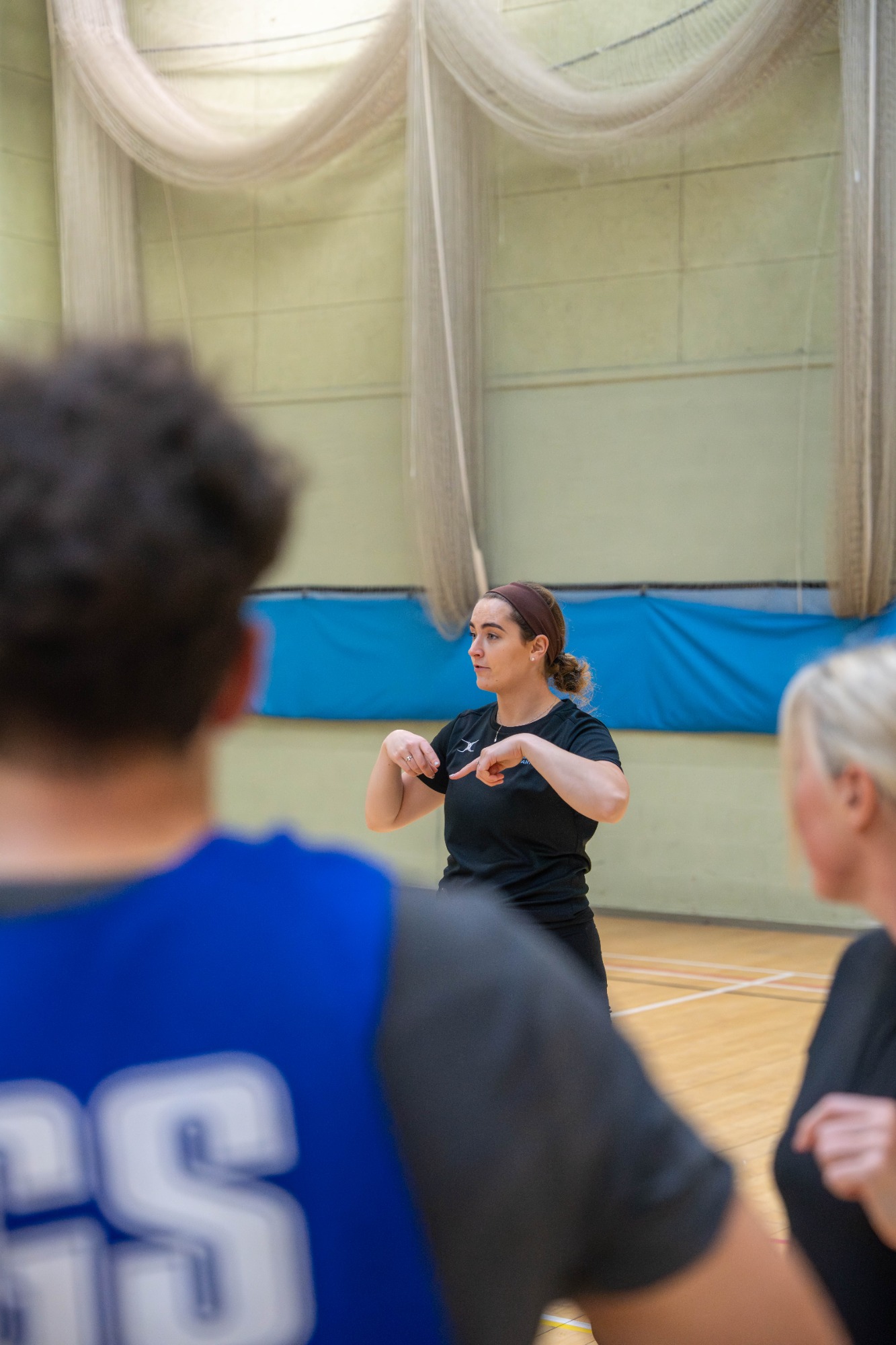 A female coach gestures while instructing players in a sports hall.