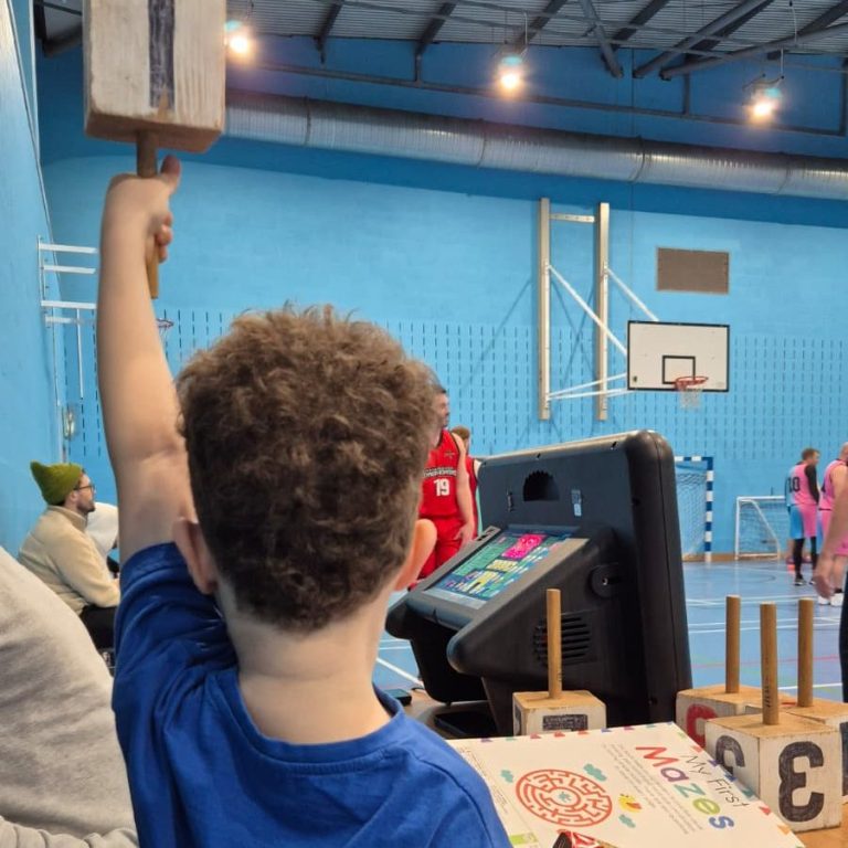 Child holding up a scoring paddle in a sports hall, with players in the background.