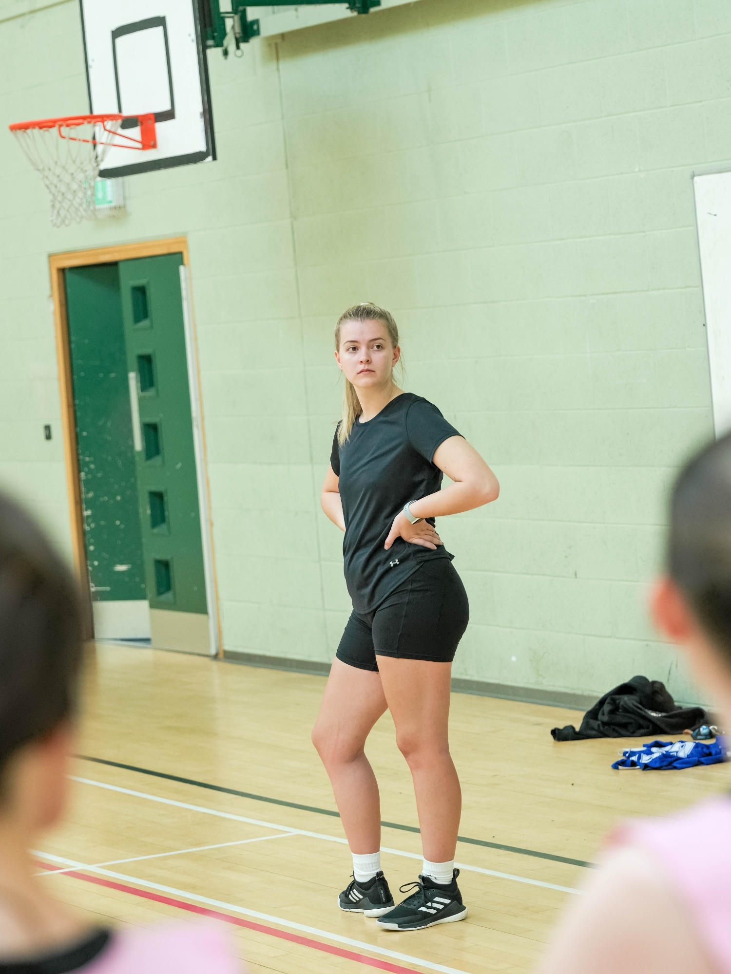 A female coach stands on a basketball court, hands on hips, speaking to players.