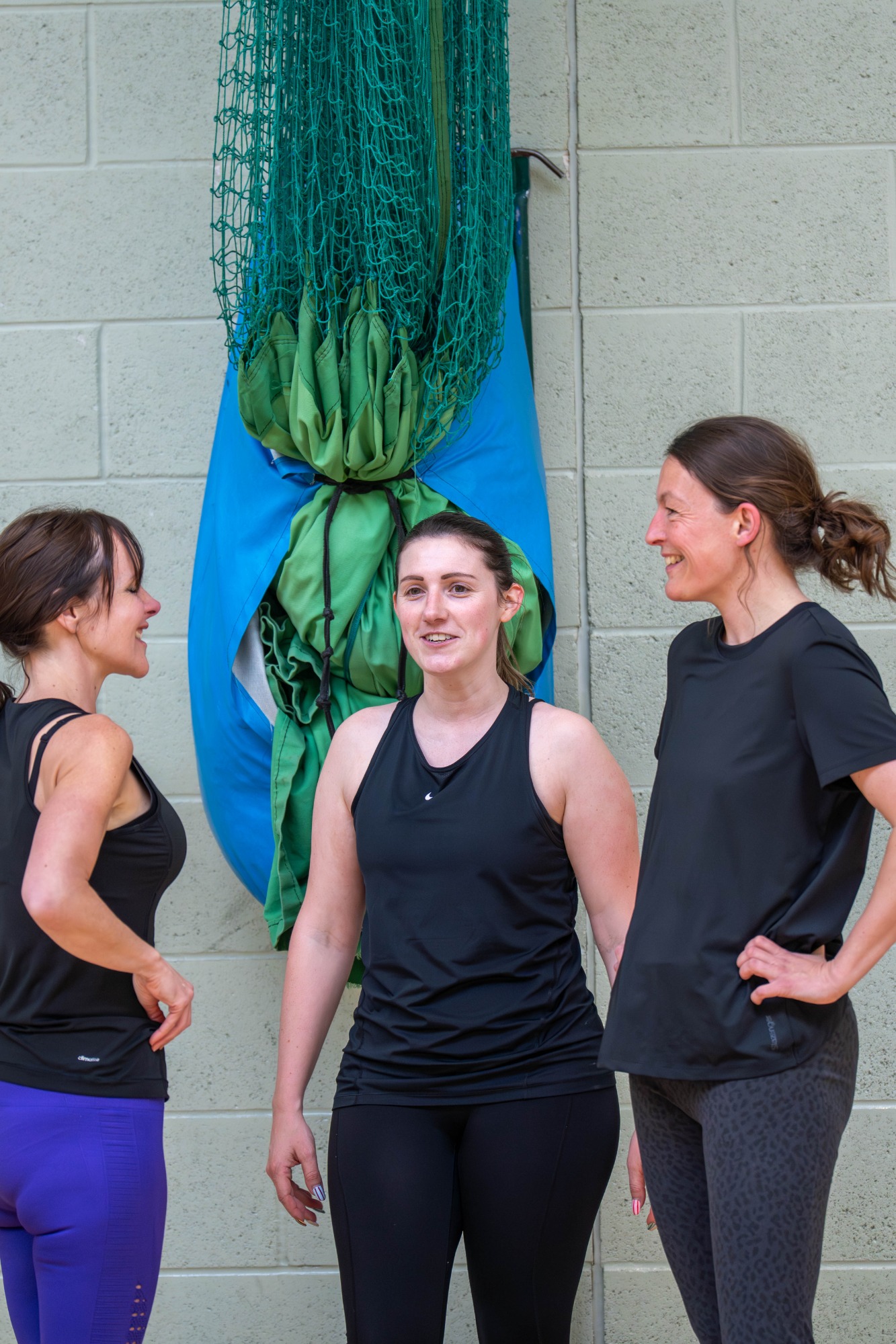 Three women in athletic clothing engage in conversation, with exercise equipment in the background.