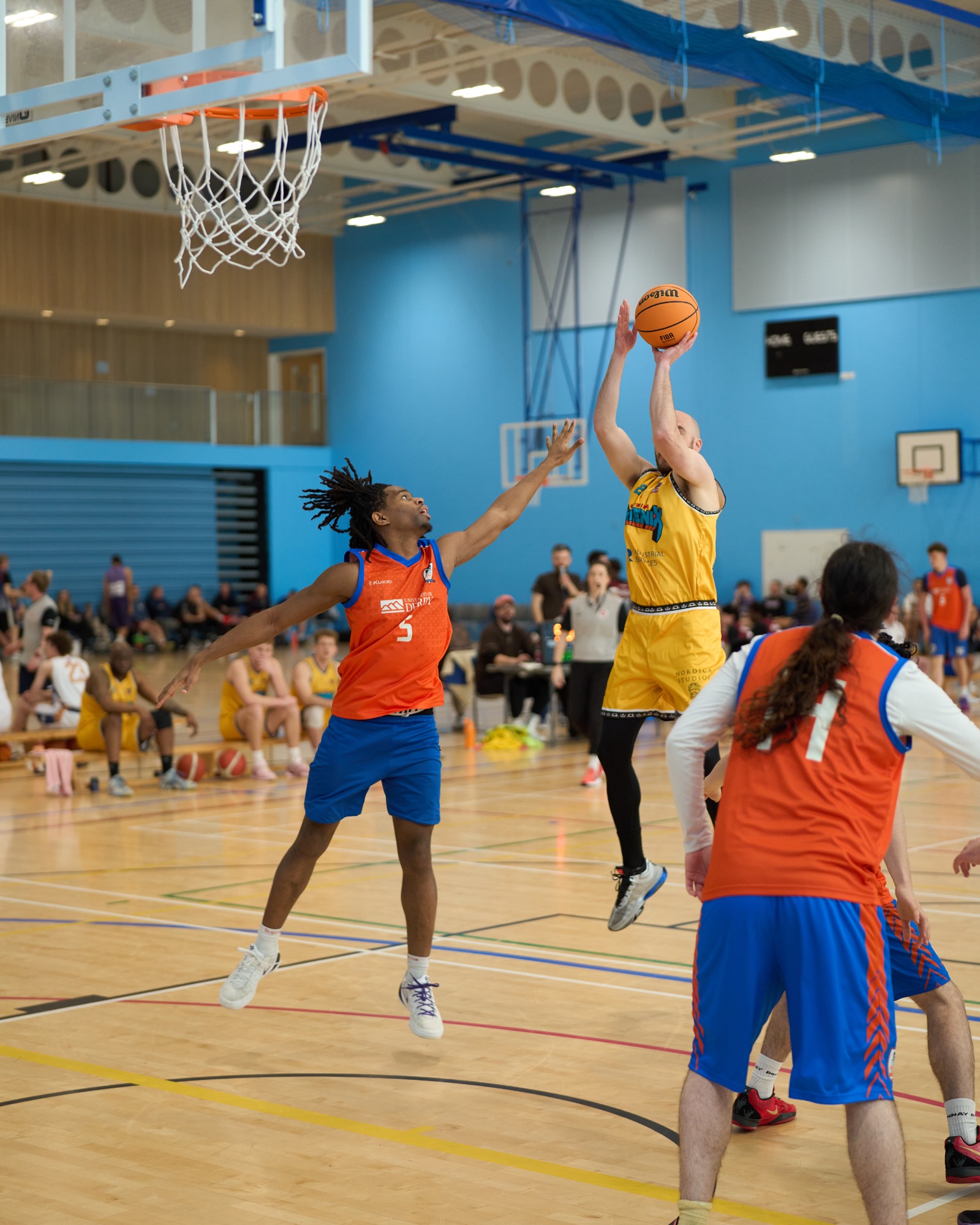 Player shoot a mid air basketball shot with player defending in mid air attempting to stop the shot going in.