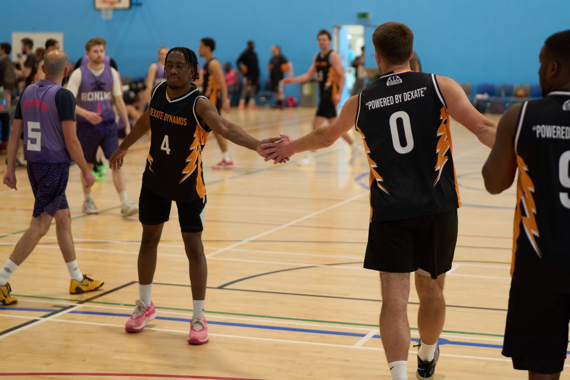 Two basketball players shake hands on the court, surrounded by teammates and spectators.