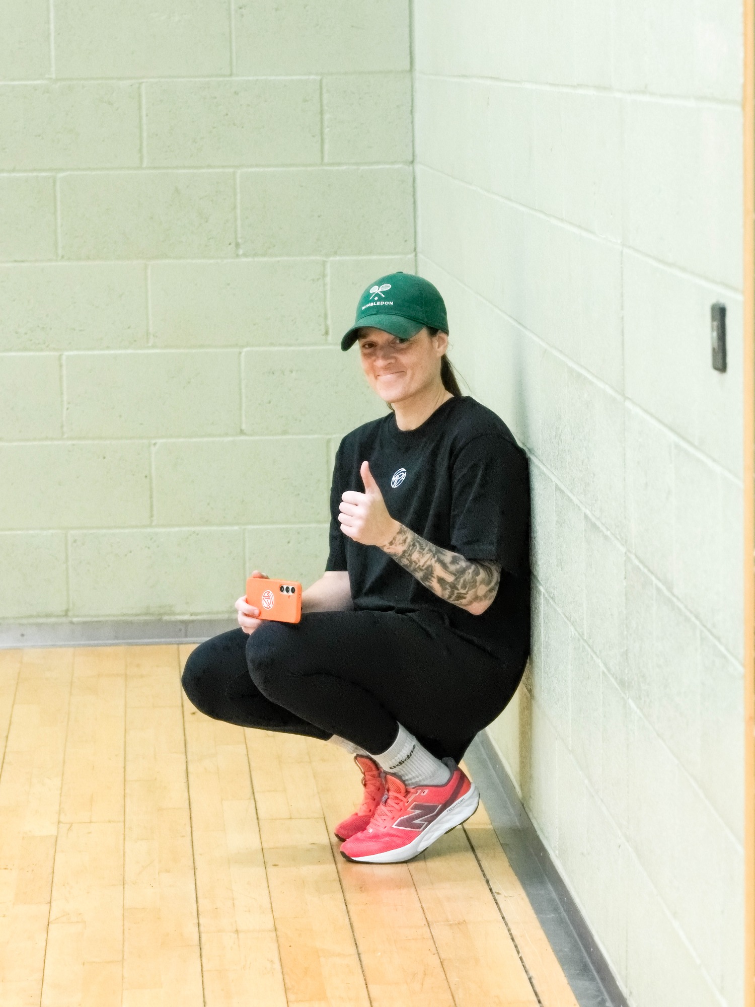 Person squatting against a wall, wearing a green cap and black outfit, giving a thumbs-up.