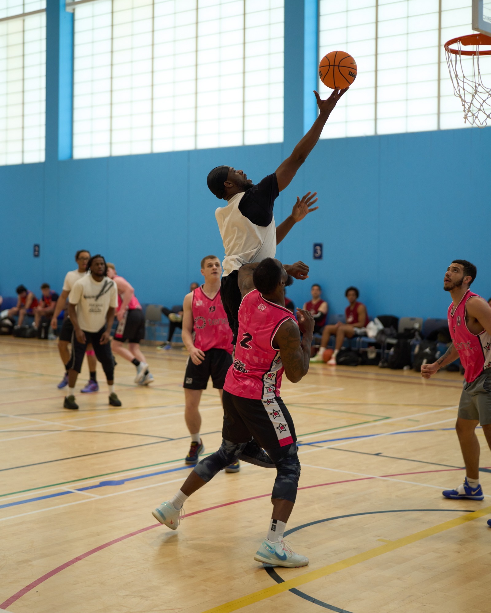 A player jumps to shoot a basketball while another defends, in a gym with spectators.
