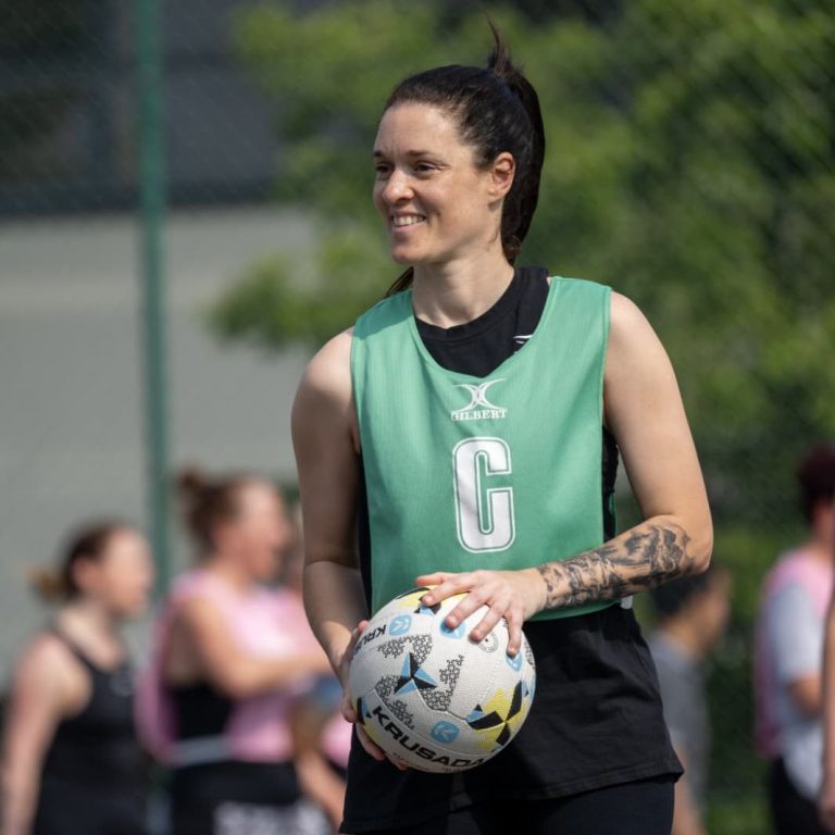 A woman in a green training vest holds a ball, smiling during a sports practice session.
