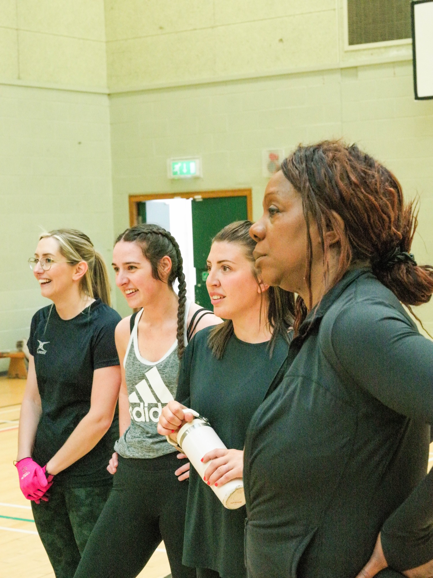 Four women in sportswear stand together, smiling and chatting in a gym setting.