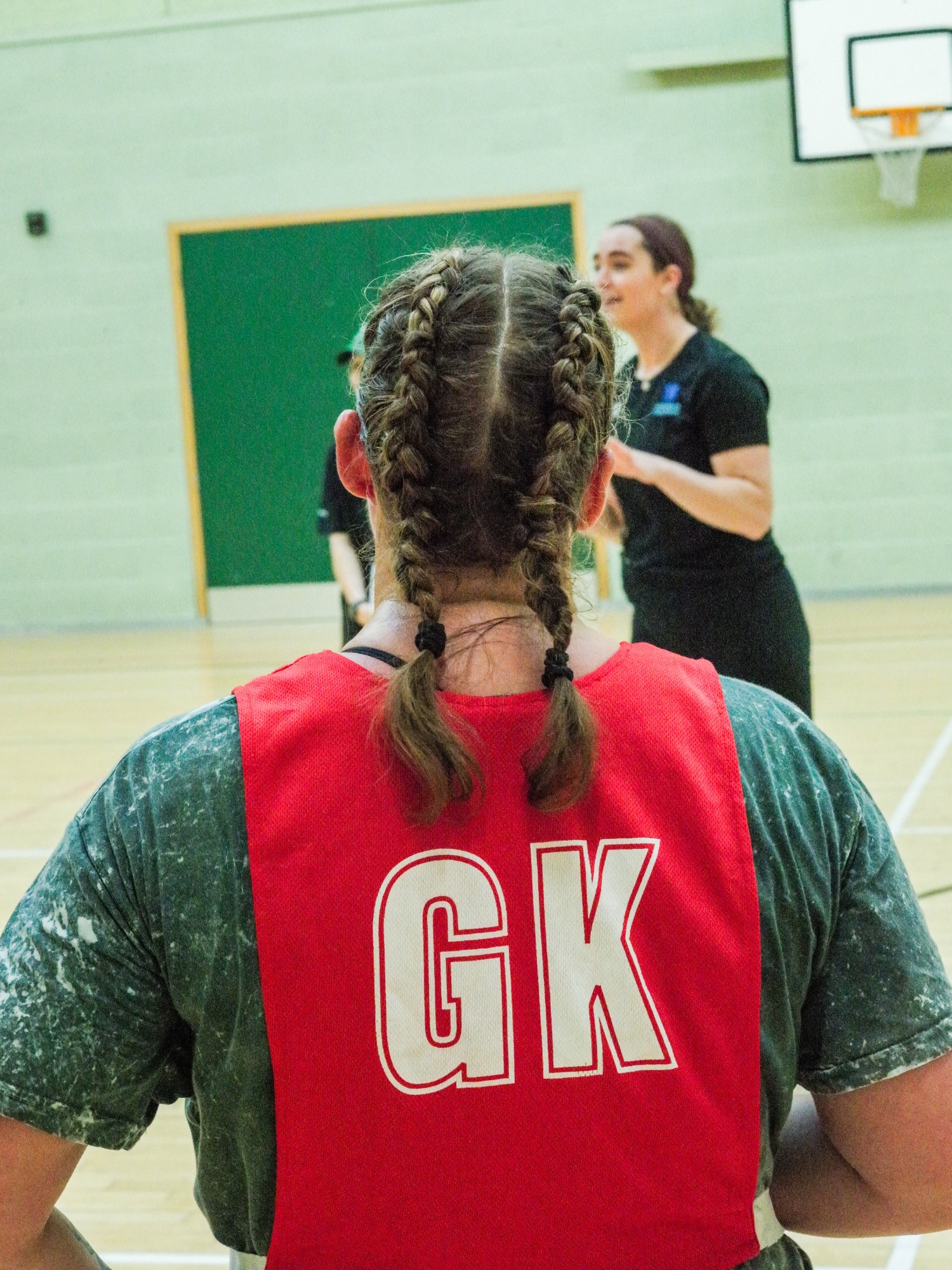 A person with braided hair wearing a red sports bib labelled "GK" watches a game.