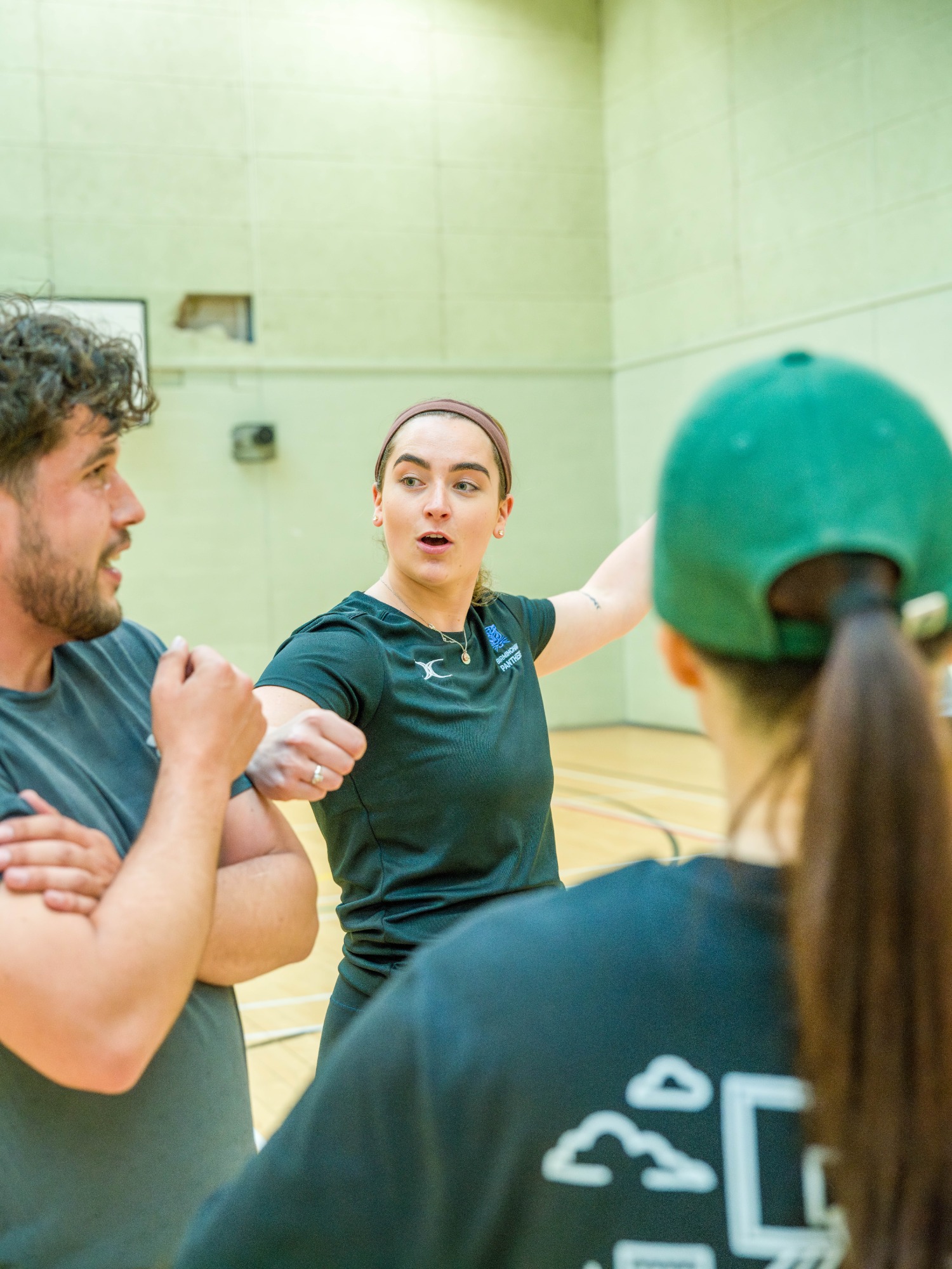 A coach gestures while discussing strategy with two athletes in a sports hall.
