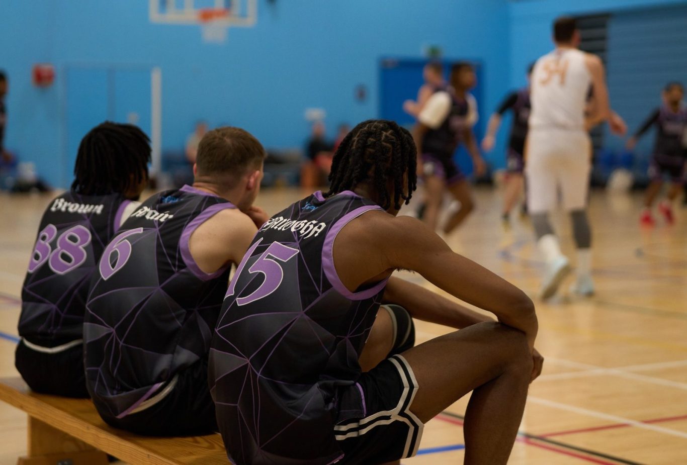Three basketball players on a bench watching a game in a gymnasium.