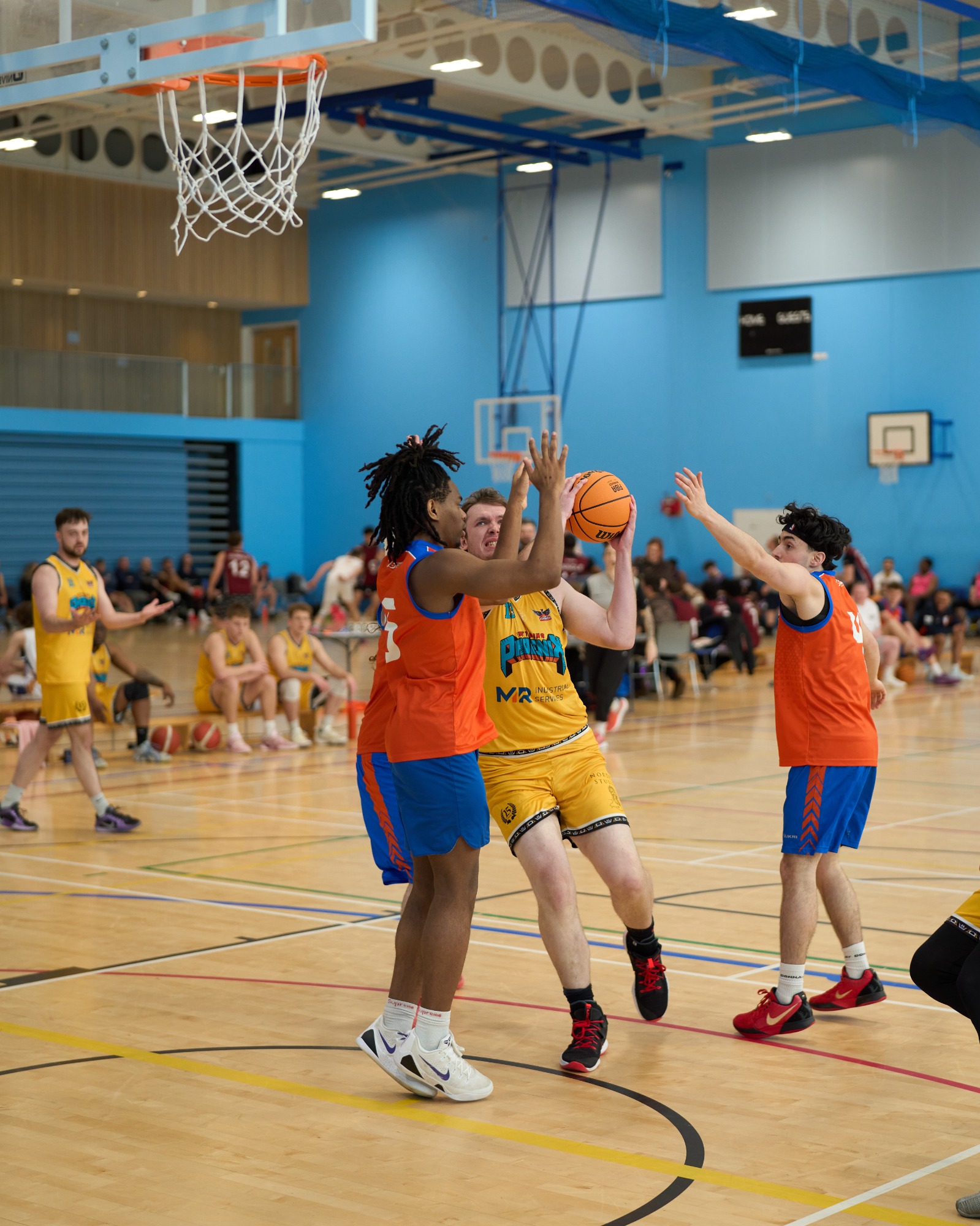 Two players in orange jerseys contest for the ball against a player in a yellow jersey.