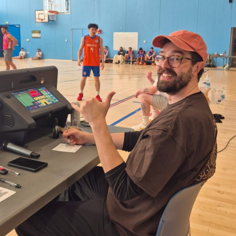 A person in a cap gives a thumbs up while sitting at a scorekeeping table in a gym.