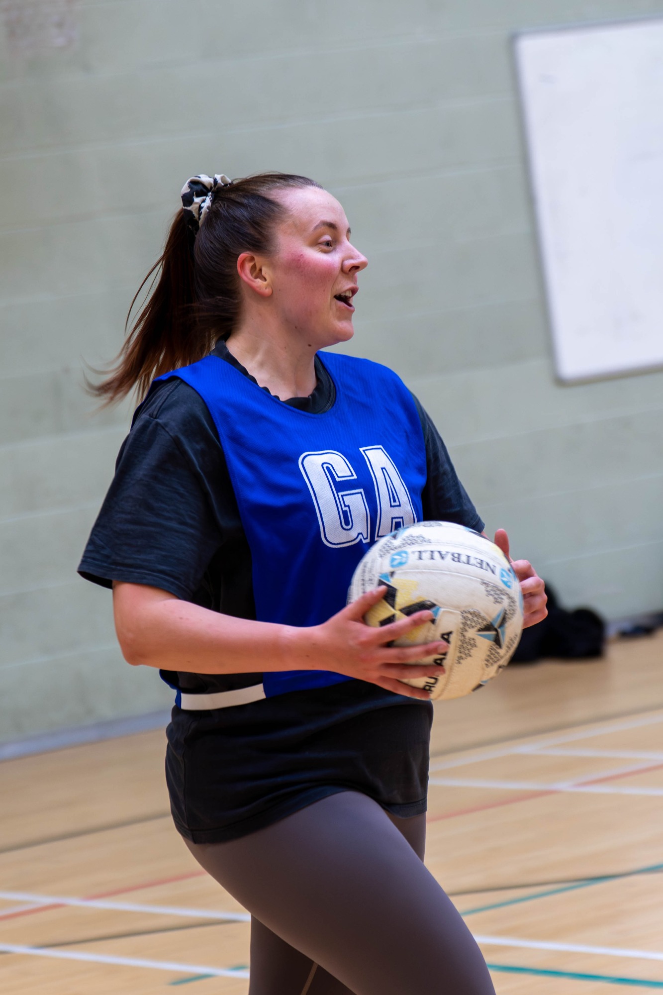 A netball player in a blue vest holds a ball, preparing to pass on a court.