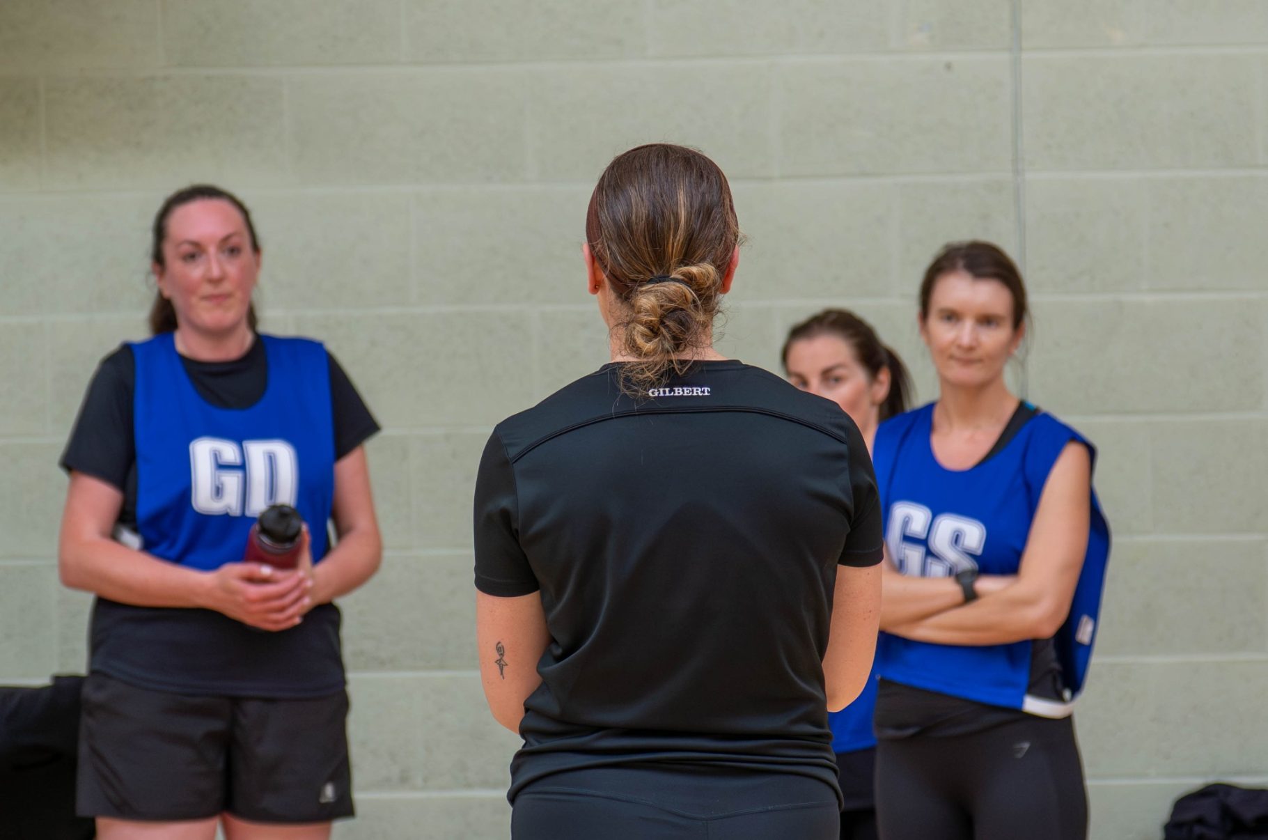 Three netball players in blue bibs watch a coach during a training session.