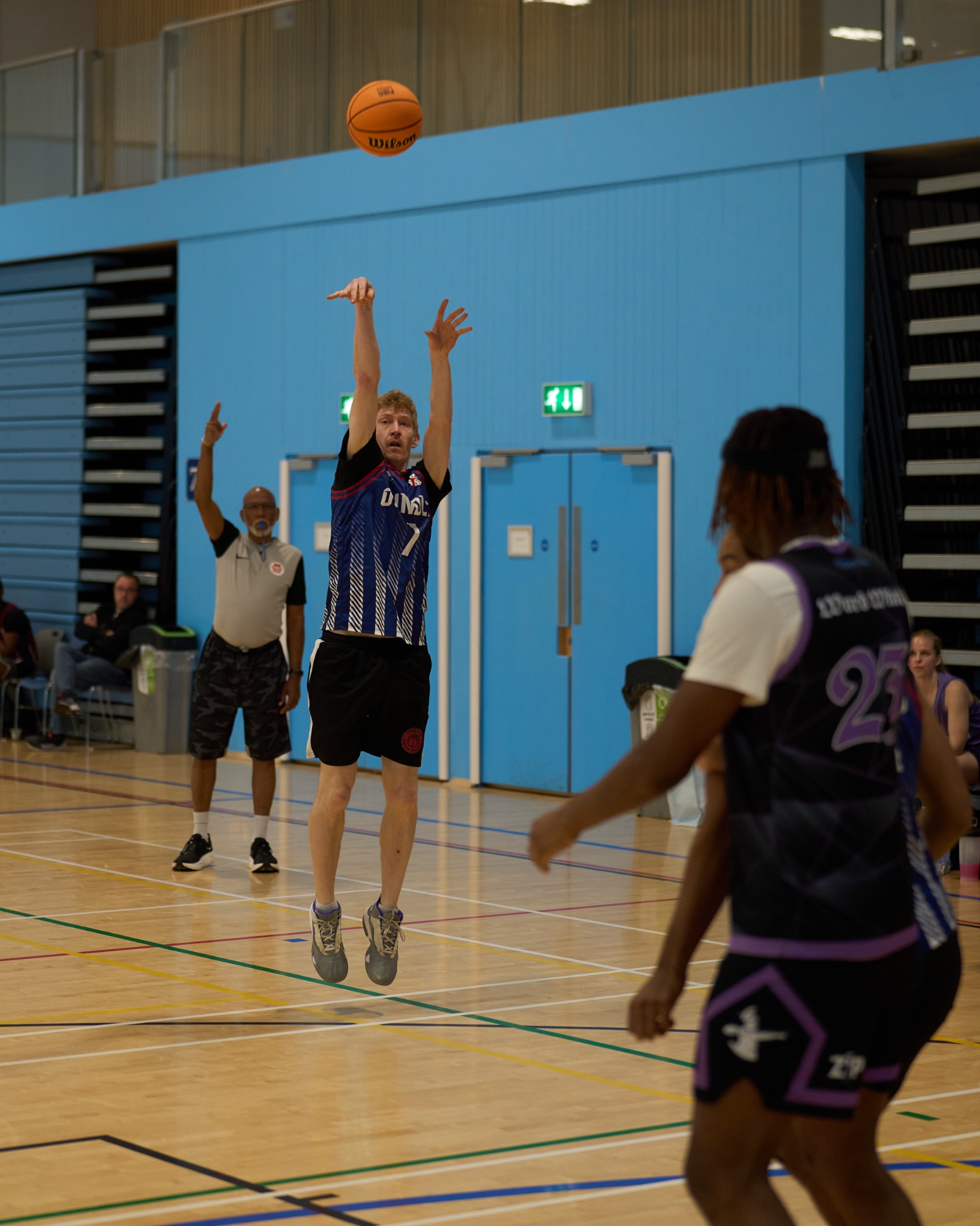 Player in mid air shooting a 3 point shot with the referee in the background signalling a 3 point attempt.