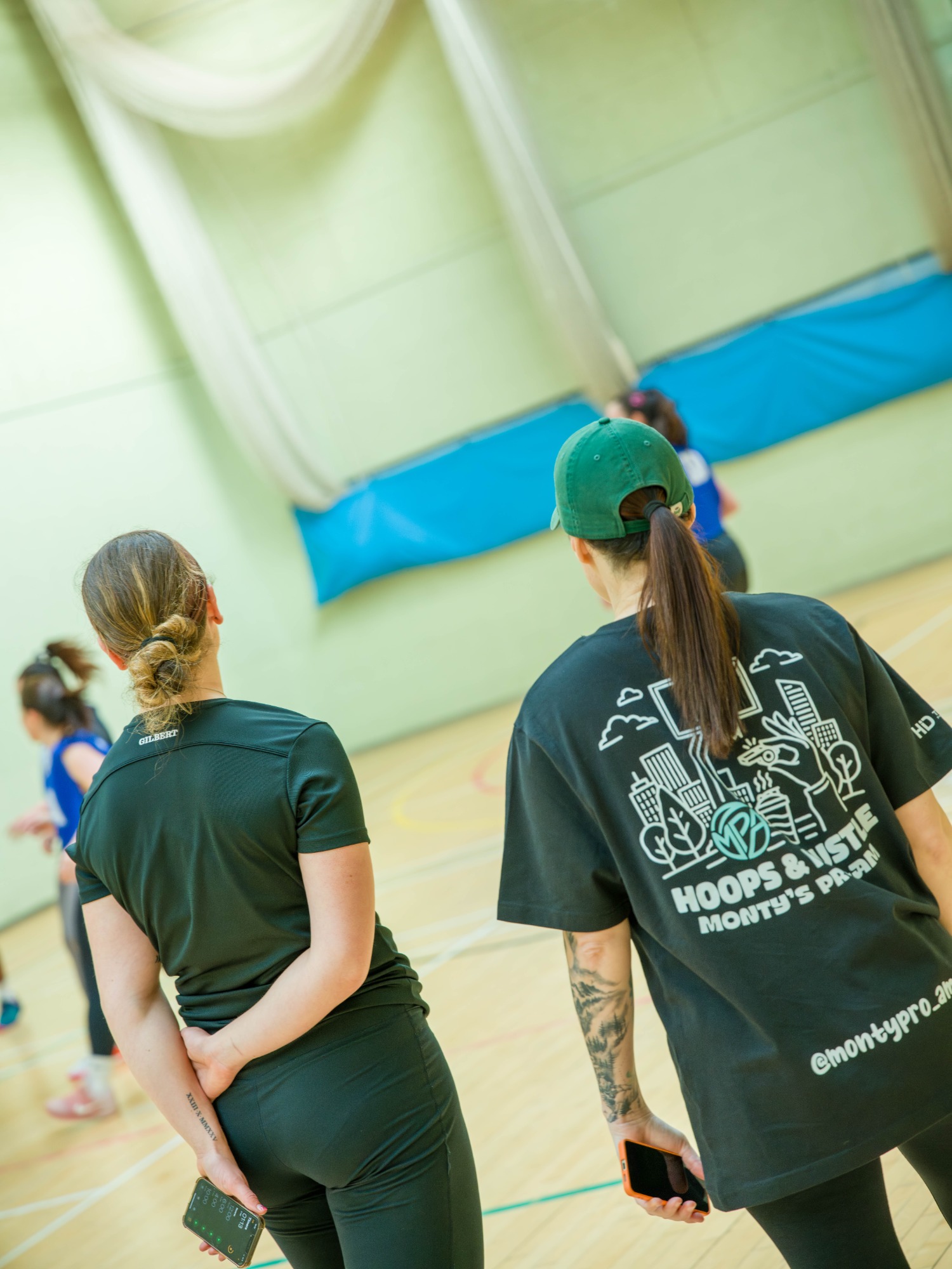 Two women observe a sports session in a gymnasium. One wears a black t-shirt and the other sportswear.