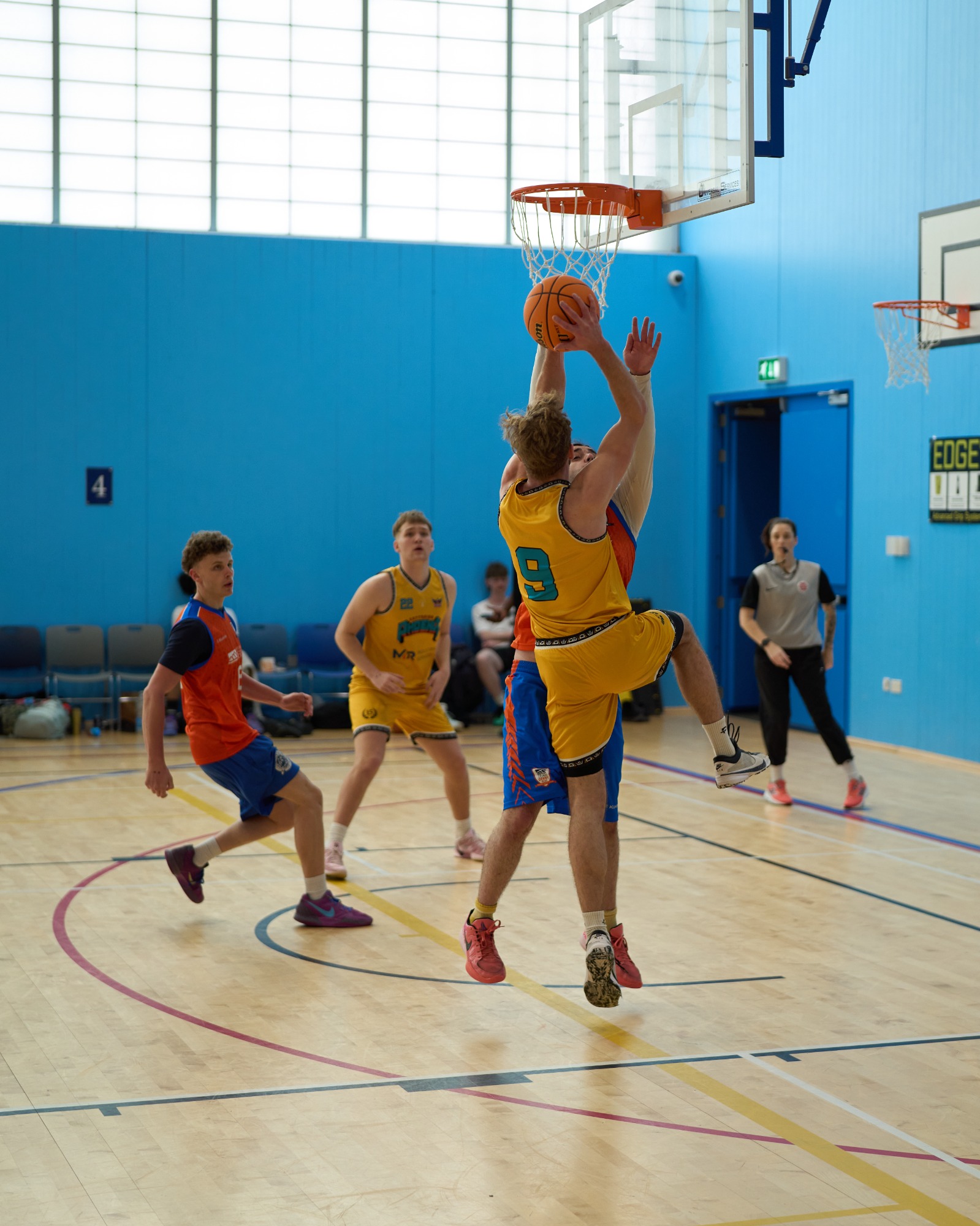 A player in a yellow jersey jumps to shoot a basketball while two opponents defend.