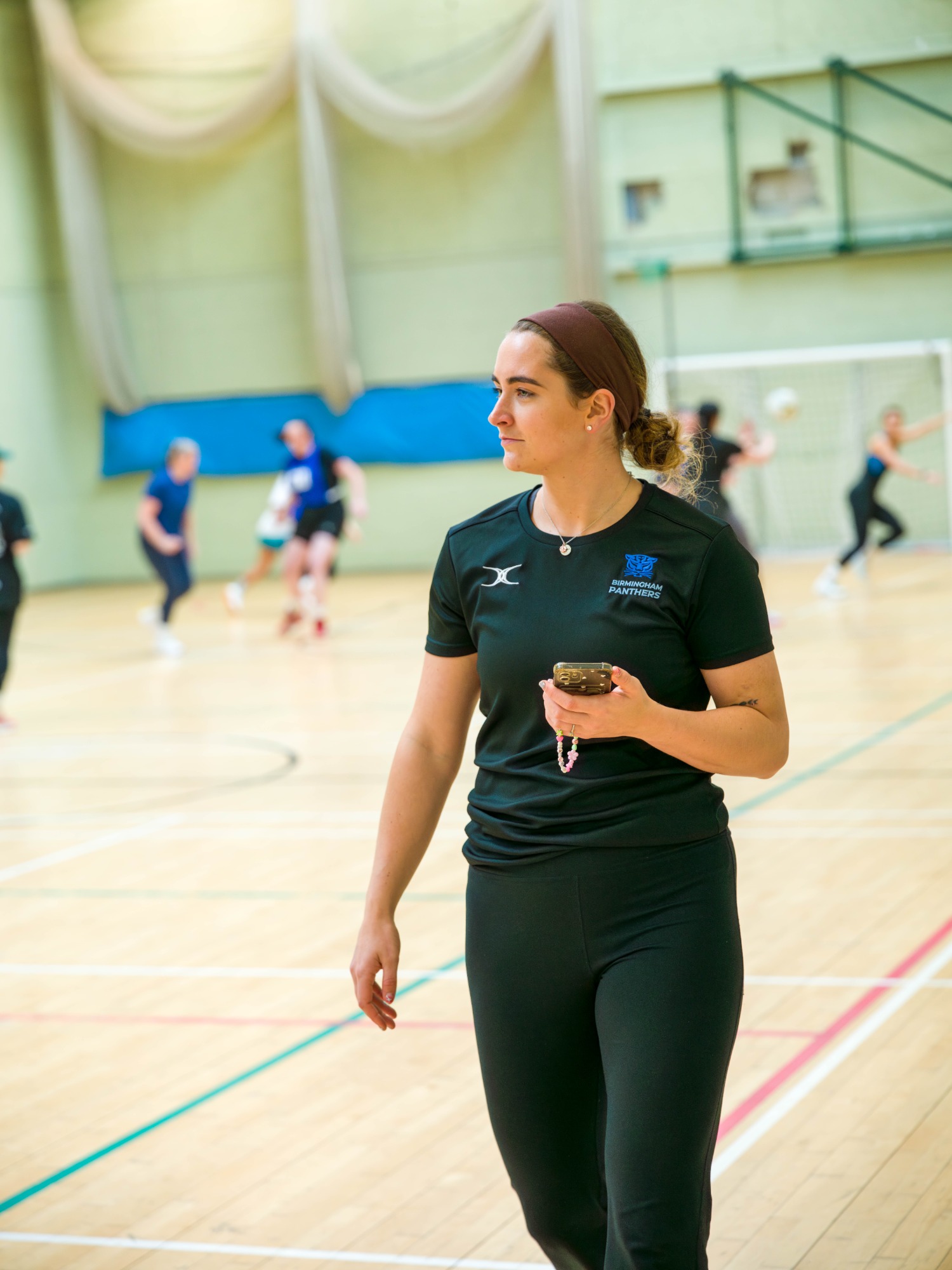A woman in a black sports shirt holds a phone while walking on a gym court.