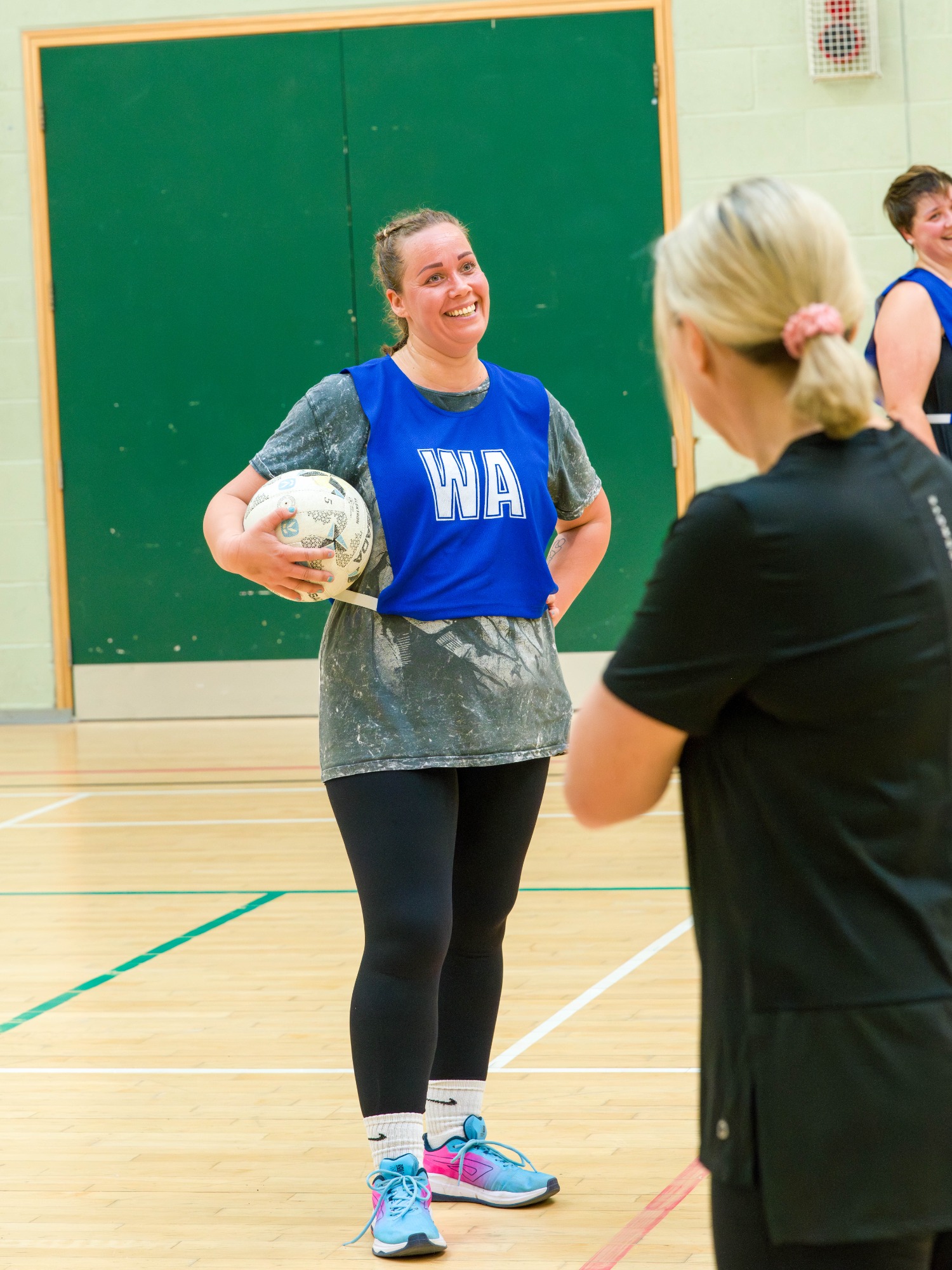 Smiling woman in sportswear holding a netball, standing in a gymnasium.